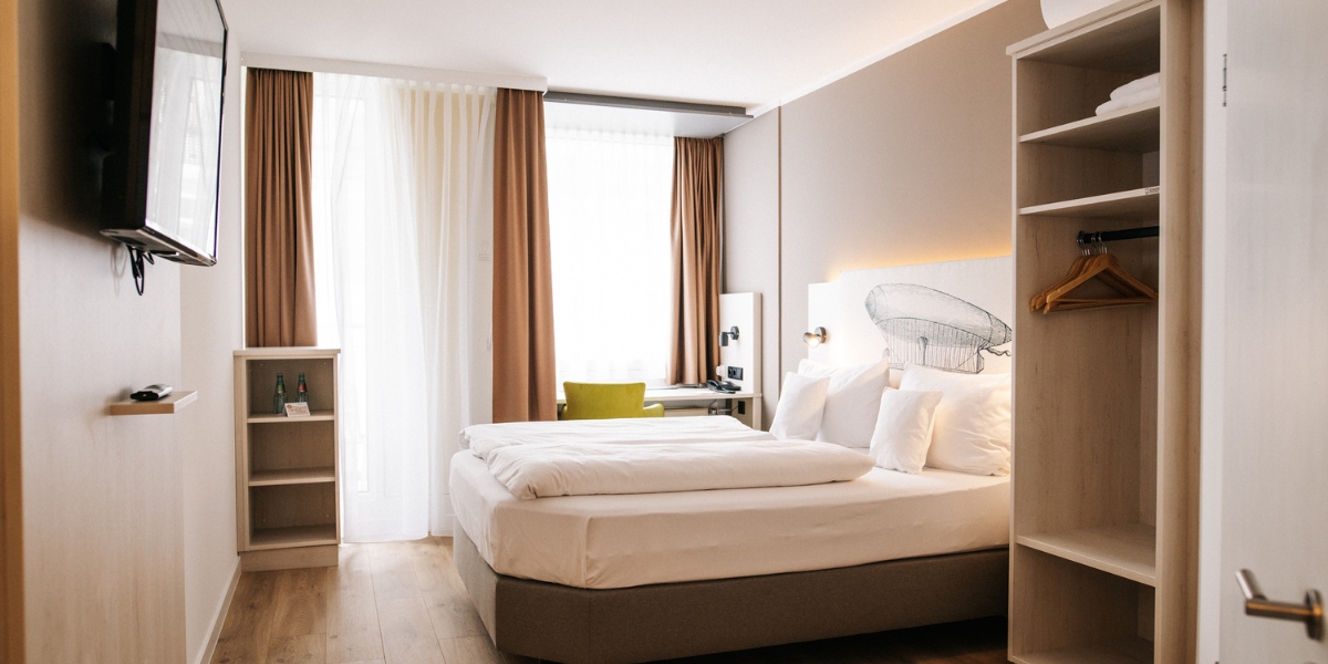 Barrierearmes Doppelzimmer im Kongresshotel Potsdam. Der Blick von der Eingangstür in das Zimmer hinein. Ein Doppelbett, ein Fernseher an der Wand, offene Schränke und ein Schreibtisch direkt vor dem Fenster.