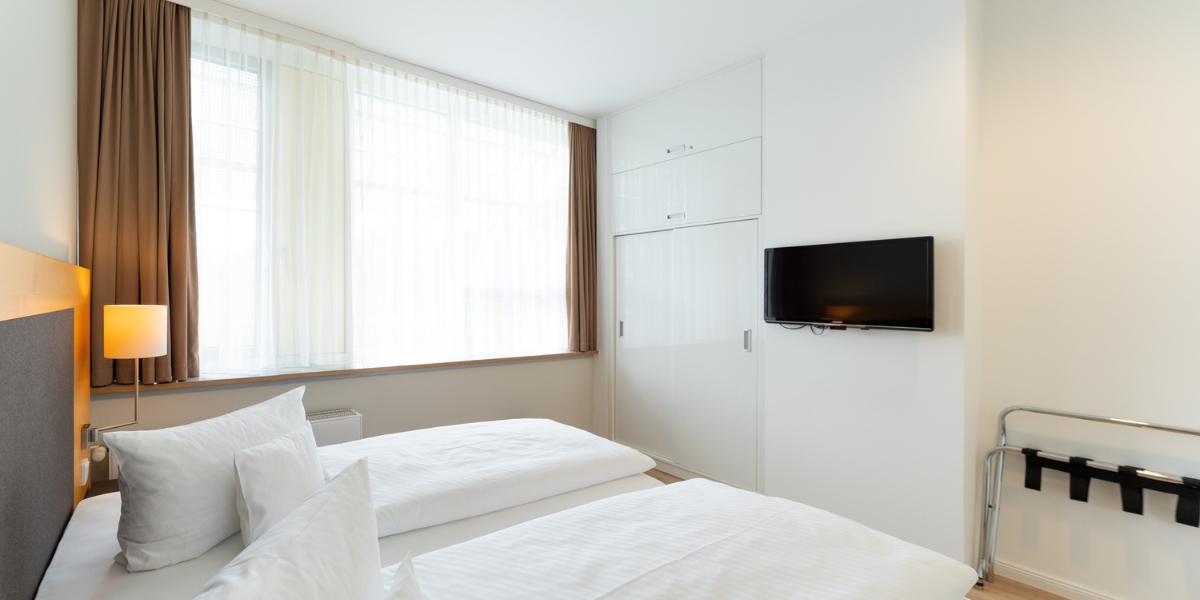 Barrierearmes Studio im Kongresshotel Potsdam. Blick von der Seite des Bettes auf das große Fenster, dem eingebauten Kleiderschrank und den Fernseher an der Wand.