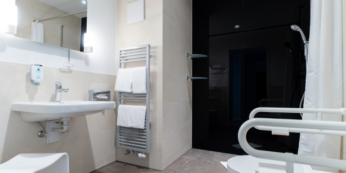 Barrierearmes Studio im Kongresshotel Potsdam. Blick auf das Badezimmer mit einem Waschbecken mit höhenverstellbaren Spiegel, einer offenen Dusche und Seitenhalterungen der Toilette.