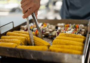 Hand mit Zange wendet Maiskolben auf Grillplatte; dahinter Gemüsespieße, Teil des Barbecue-Buffets