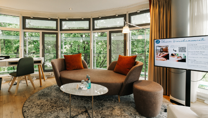 Heller Wohnbereich mit großer Fensterfront, Sofa mit Kissen, kleinem Tisch mit Wasserflaschen sowie Schreibtisch und Bildschirm mit Wellnessangeboten.