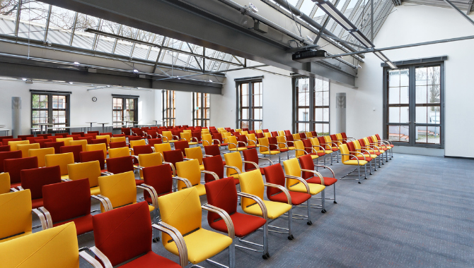 Moderner Tagungsraum mit großen Fenstern und Reihen aus rot-gelben Stühlen.
