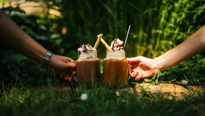 Zwei Hände stoßen mit dekorierten Milkshakes im Grünen an.