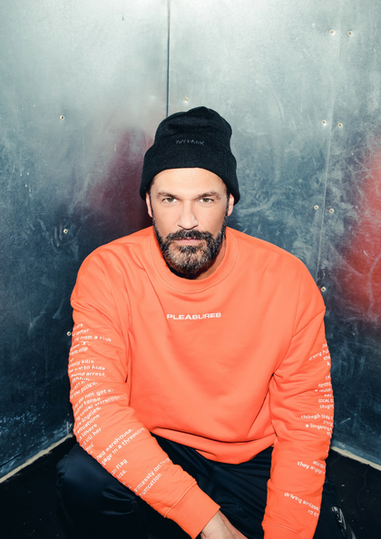 Mann mit orangefarbenem Sweatshirt sitzt vor metallischer Wand.