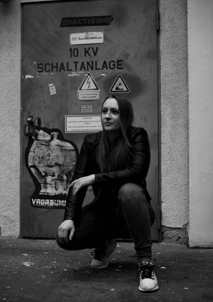 Frau in Lederjacke hockt vor einer Tür mit Warnhinweisen in Schwarz-Weiß-Aufnahme.