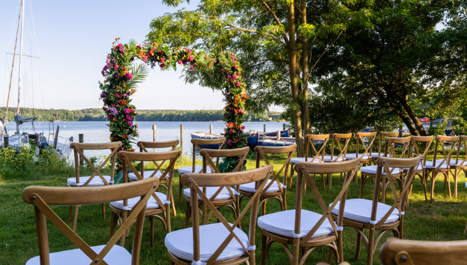 Freie Trauung am See mit Holzstühlen und floral geschmücktem Traubogen.