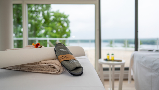 Kongresshotel-Potsdam-Seeblick-Sauna (2) Massageliege mit Handtüchern und Hausschuhen in einem hellen Wellnessraum. Große Panoramafenster bieten einen weiten Blick auf den See.