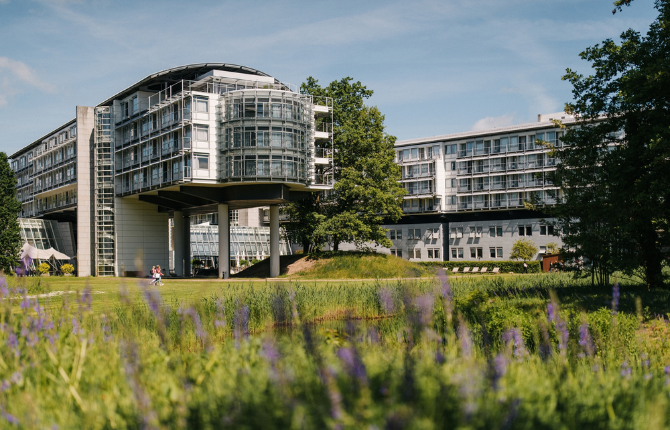 Kongresshotel-Potsdam–Außenansicht–Sommer Außenansicht des Kongresshotels Potsdam mit markanter Architektur, umgeben von weitläufigen Grünflächen und blühenden Wiesen in sommerlicher Atmosphäre.
