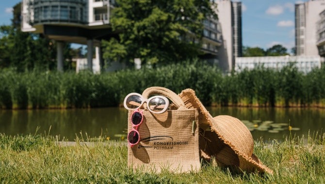 Kongresshotel-Potsdam–Urlaubswoche Strandtasche mit Sonnenbrillen und Sonnenhut auf einer Wiese am Wasser vor dem Kongresshotel Potsdam. Die Szene vermittelt entspannte Urlaubstage in naturnaher Umgebung.